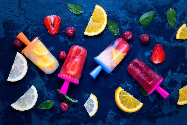 Ghiacciolo di frutta brillante fatto in casa con aroma di fragola, ciliegia, limone, arancia, limone e menta e frutta fresca per gelato su un blu scuro