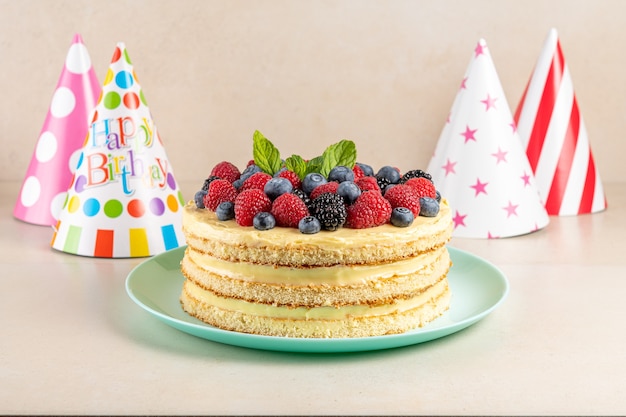 Homemade cake with fresh berries and birthday hat on bright surface.