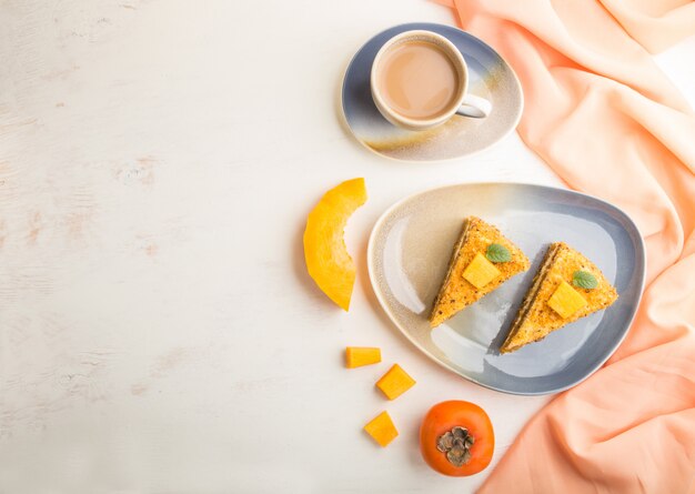 Homemade cake with persimmon and pumpkin and a cup of coffee on a white wooden background with orange textile. top view, copy space.