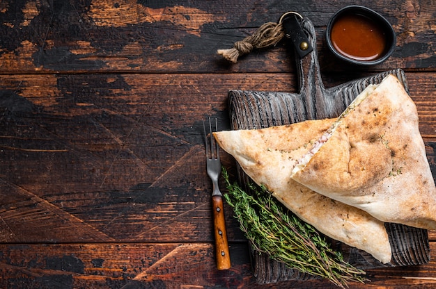 Homemade Calzone closed pizza with ham and cheese on wooden board. Dark wooden background. Top view. Copy space.