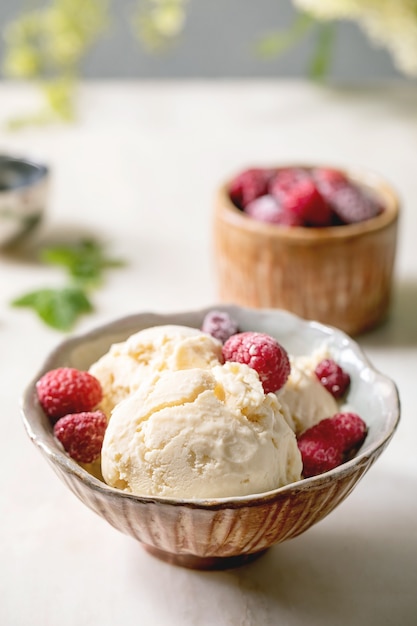 Homemade caramel vanilla ice cream with frozen raspberries in ceramic bowl standing on white marble table with flowers behind