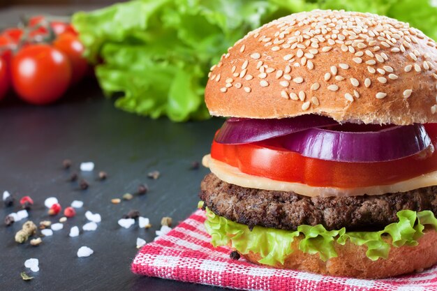 Homemade cheeseburger with beef patties, fresh salad, tomatoes and onion on seasame buns, served on black slate table.