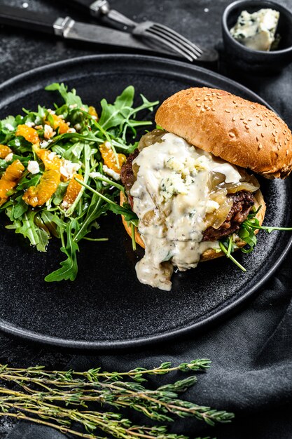 Homemade cheeseburger with blue cheese, bacon, marbled beef and onion marmalade, a side dish of salad with arugula and oranges. Black surface. Top view