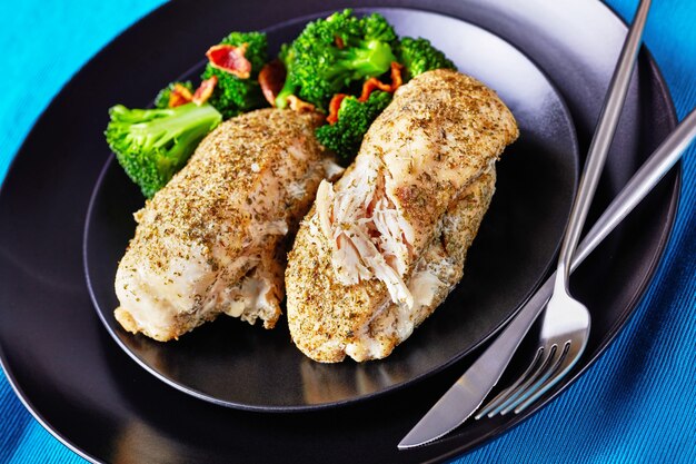 Foto filetto di pollo fatto in casa al forno con condimento ranch servito su un piatto nero con broccoli e pancetta con posate d'argento su uno sfondo di cemento scuro, orientamento orizzontale, primo piano