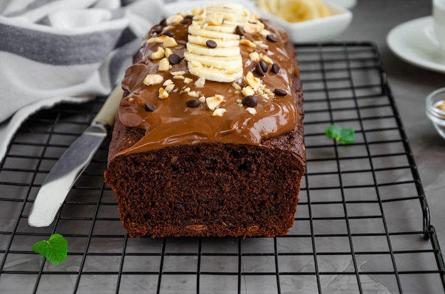 Homemade chocolate banana bread with chocolate cream, banana slices and nuts on top on a dark concrete background. Horizontal orientation.