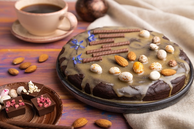 Homemade chocolate brownie cake with caramel cream and almonds with cup of coffee on a colored wooden background. 