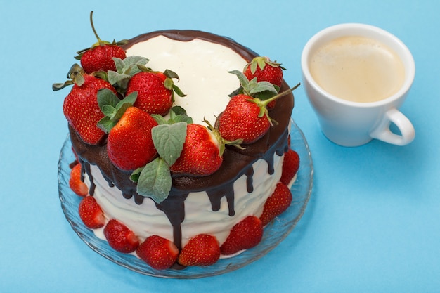 Homemade chocolate cake decorated with fresh strawberries and leaves of mint on glass plate and cup of coffee on blue color background