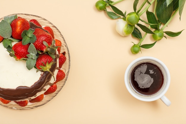 Homemade chocolate cake decorated with fresh strawberries and leaves of mint on glass plate, cup of coffee and bouquet of peonies on beige color background. Top view