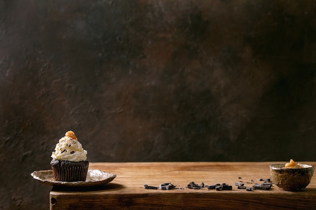 Homemade chocolate cupcakes muffins with white whipped butter cream and salted caramel on ceramic plate, served with chopped dark chocolate on wooden table. Copy space