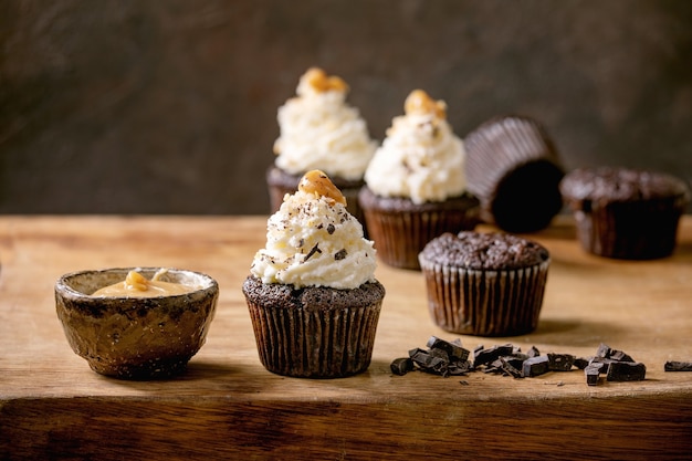 Homemade chocolate cupcakes muffins with white whipped butter cream and salted caramel on ceramic plate, served with chopped dark chocolate on wooden table.