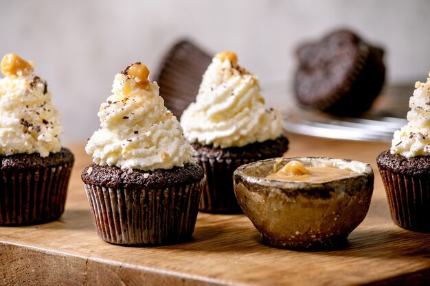 Домашние шоколадные кексы кексов с белым взбитым сливочным кремом и соленой карамелью на керамической тарелке на деревянном столе.