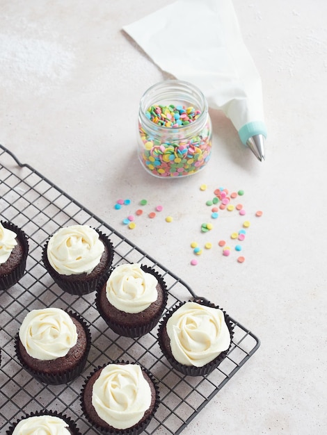 Homemade chocolate cupcakes with white buttercream on cooling rack Piping bag with starshaped nozzle and colored edible confetti for decorating pastries