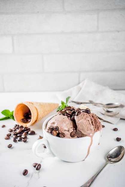 Homemade coffee ice cream, served with coffee beans and mint leaves, with ice cream cones and spoons in the picture. White marble background,