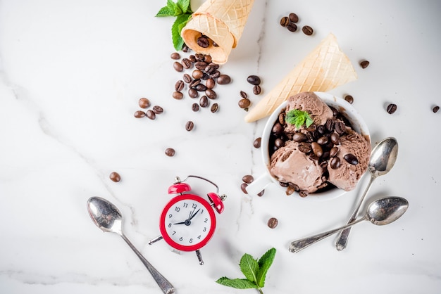 Homemade coffee ice cream, served with coffee beans and mint leaves, with ice cream cones and spoons in the picture. White marble background,