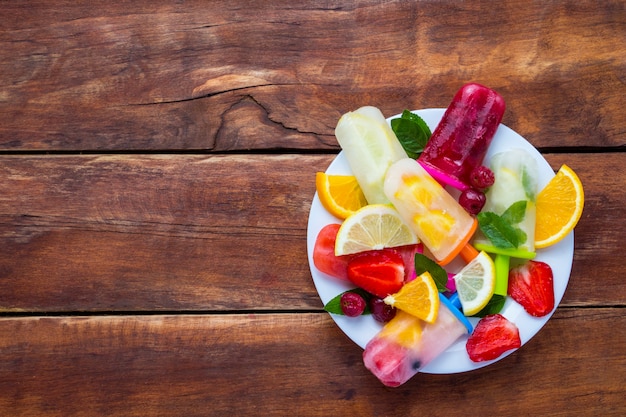 Homemade colorful, fruit popsicle, Fresh fruit on a white plate and a dark wooden background. Strawberry, Lemon, Lemon with mint, Orange, Cherry, Multifruit. Flat lay, top view