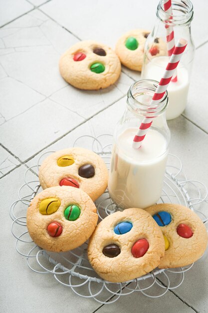 Homemade cookies with colorful chocolate candies and milk Stack of shortbread cookies with multi colored candy on plate with bottle of milk on light gray background Baby food concept Copy space