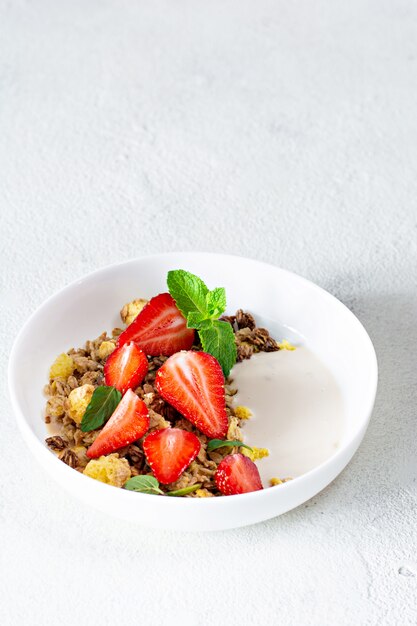 Homemade crunchy granola with nuts, dried fruits, fresh strawberries, mint and yogurt (fermented baked milk) on a white plate.