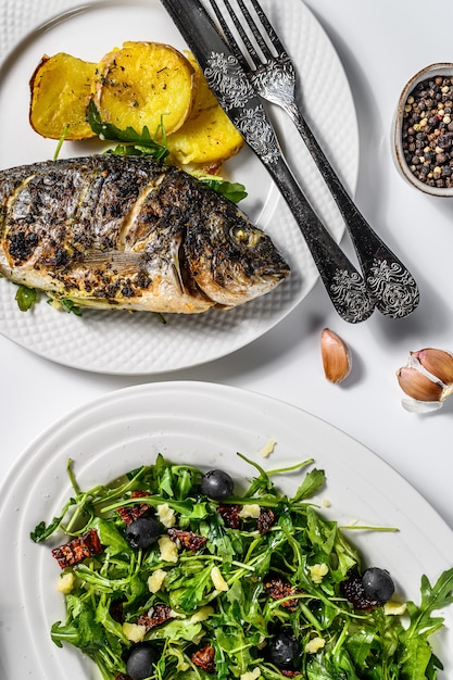 Homemade dinner with baked Dorado fish, arugula salad with tomatoes, baked potatoes. White background. top view