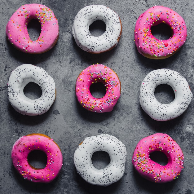 Homemade doughnuts pink with rainbow sprinkles and white with icing poppy seeds on the dark grey concrete background. Free copy space. Top view. Abstract pattern. Square crop