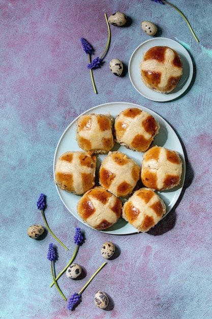 Homemade Easter traditional hot cross buns on ceramic plate with muscari flowers, quail eggs, textile over blue texture surface