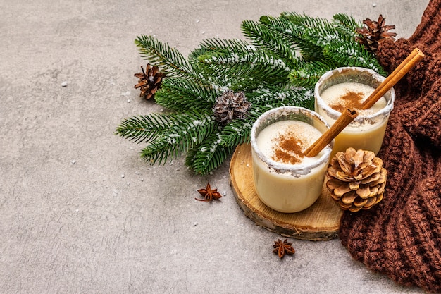 Домашнее eggnog с корицей в стекле. Типичный рождественский десерт. Вечнозеленые еловые ветки, шишки, уютный плед, искусственный снег.