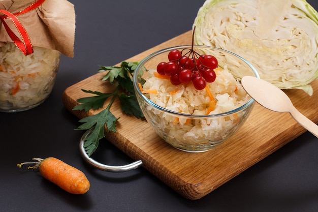 Homemade fermented cabbage with carrot in glass bowl decorated with cluster of viburnum. Fresh head of cabbage and glass jar on the background. Vegan salad. Dish is rich in vitamin U.