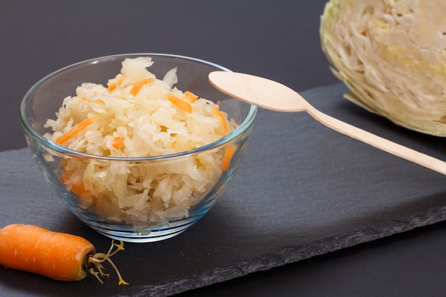 Homemade fermented cabbage with carrot in glass bowl with fresh head of cabbage on black background. Vegan salad. Dish is rich in vitamin U. Food great for good health.