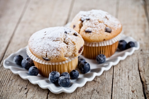 Homemade fresh muffins with sugar powder and blueberries on ceramic plate on rustic wooden table.