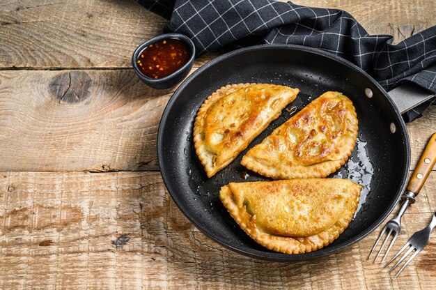 Фото Домашние жареные чебуреки с мясом и зеленью на сковороде, традиционная кавказская кухня. деревянный фон. вид сверху. скопируйте пространство.