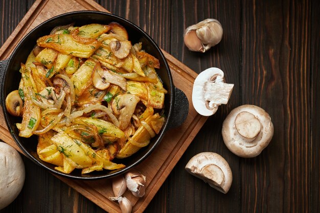 Foto patate fritte fatte in casa, con cipolle, funghi e aglio, in padella, su una tavola di legno