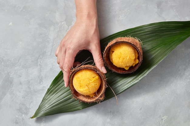 Homemade fruit ice cream in a coconut shell on a palm leaf on a gray concrete table. A girl's hand takes a shell with ice cream. Copy space for your text. Top view