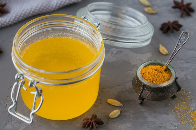 Ghee fatto in casa o burro chiarificato in un barattolo e polvere di curcuma su cemento grigio.