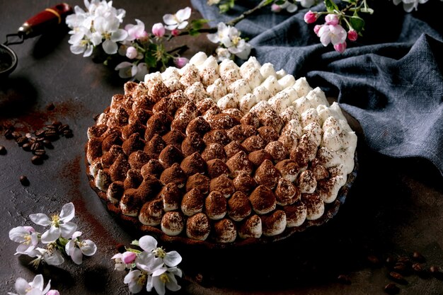 Homemade gluten free tiramisu traditional italian dessert sprinkled with cocoa powder decorated with blooming apple tree, blue textile napkin and coffee beans over dark texture surface.