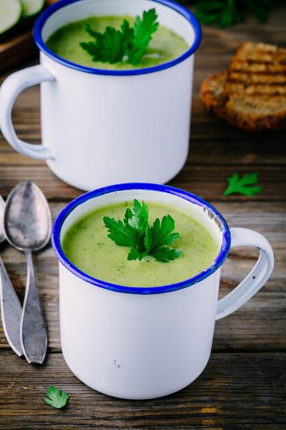 Homemade green broccoli cream soup with parsley in mugs on wooden background