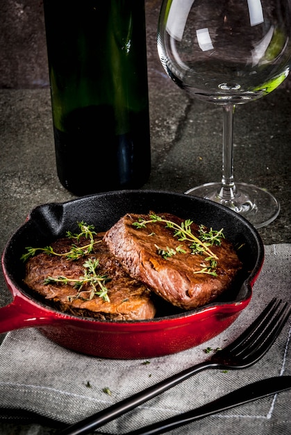 Homemade grilled beef steaks in a portioned frying pan, with a fork, knife and a glass of wine on black stone table