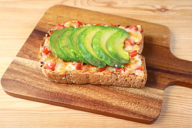 Homemade grilled cheese toast topped with tomato and sliced avocado on wooden Breadboard