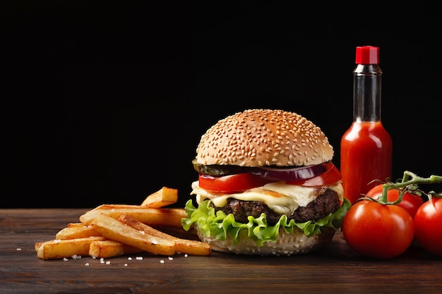 Крупный план домашнего гамбургера с говядиной, помидорами, салатом, сыром, луком, картофелем фри и бутылкой соуса на деревянном столе. Фастфуд на темном фоне.