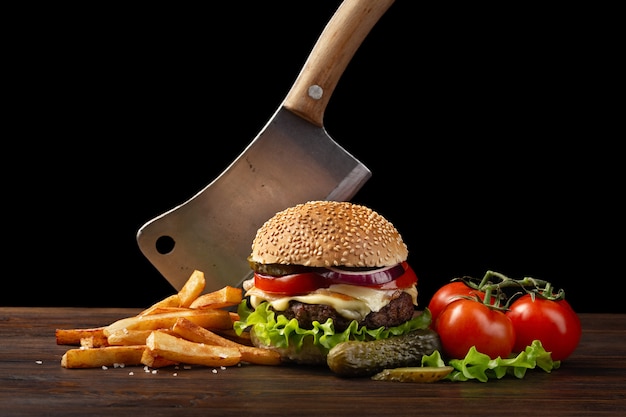 Homemade hamburger close-up with beef, tomato, lettuce, cheese, onion and french fries on wooden table. In the burger stuck a knife. Fastfood on dark background