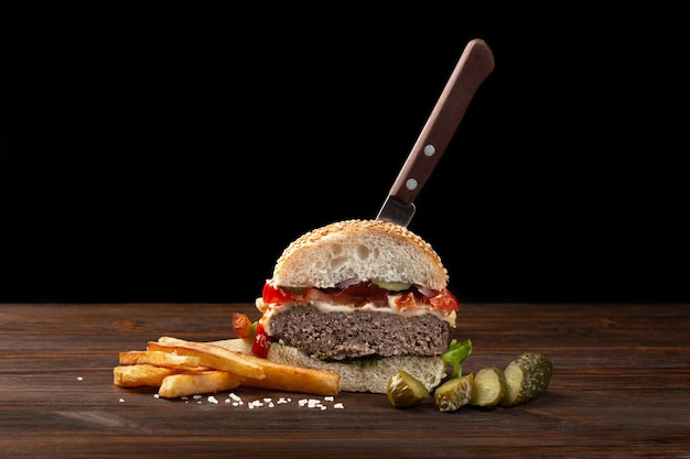 Домашний гамбургер, разрезанный пополам крупным планом с говядиной, помидорами, салатом, сыром и картофелем на деревянном столе. В бургер воткнули нож. Фастфуд на темном фоне.