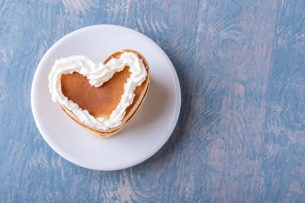 Homemade heart shaped pancake decorated with white cream on a white plate on a blue wooden background