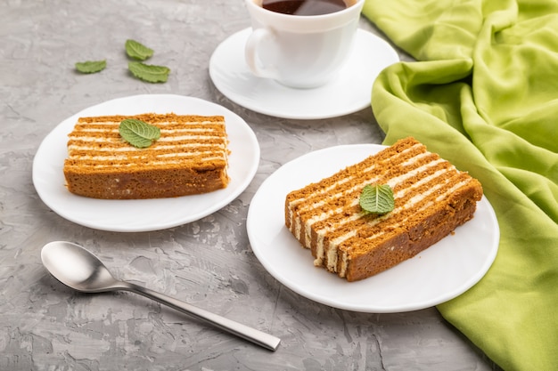 Homemade honey cake with milk cream and mint with cup of coffee on a gray concrete background. Side view, close up.