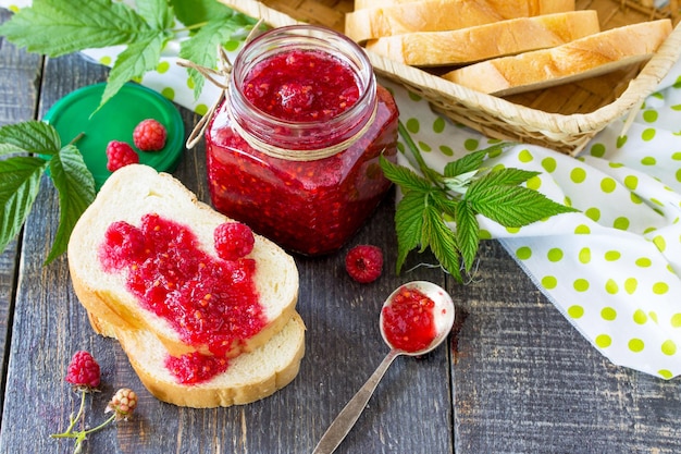 Homemade jam Glass jar with red raspberries and white bread on the kitchen table Preserved berry