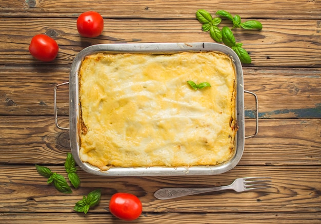 Homemade lasagna ready to eat in metal baking tray served with basil leaves and tomatoes on the brown wooden background. Top view.