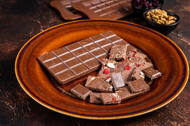 Homemade milk chocolate with hazelnuts, peanuts, cranberries and freeze dried raspberries on a rustic plate. Dark background. Top view.