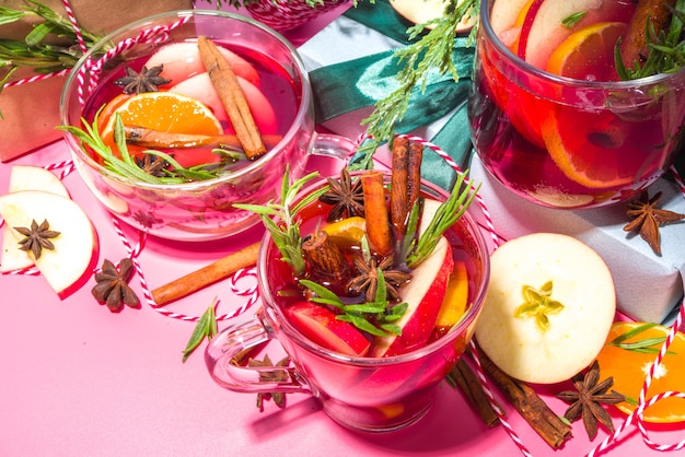 Foto cocktail di vin brulè fatto in casa. varie tazze di vetro con classico vin brulè caldo e piccante invernale, con ingredienti, decorazioni natalizie e scatole regalo su sfondo moderno ad alto colore