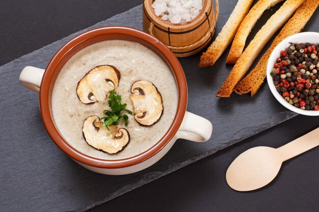 homemade mushroom cream soup with sliced mushrooms in porcelain bowl, wooden spoon, allspices, toasts and little wooden barrel with salt on a black stone board. top view.