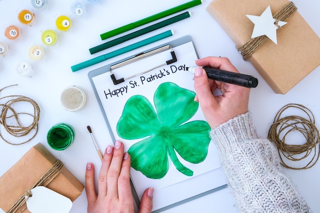 Homemade painted greeting card St Patricks day. Gift idea, decor. DIY. Do it yourself. Clovers, brush and paint. Female hands. Saint Patricks Day hat.