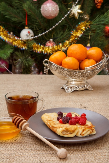 Homemade pancakes on plate, cup of tea and honey in glass bowl with wooden spoon, metal vase with oranges on sackcloth. Fir tree with toy balls and garland on the background.