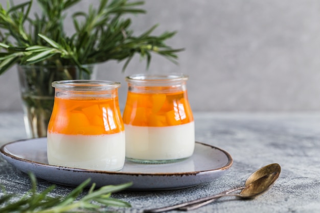 Homemade panna cotta with slices of peach and peach jelly in glass jars on a gray concrete background