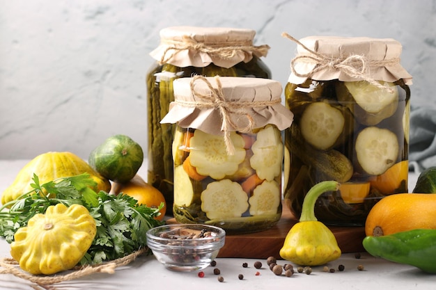 Homemade pickled patissons cucumbers and zucchini in glass jars and fresh ingredients on a light gray background in still life Closeup